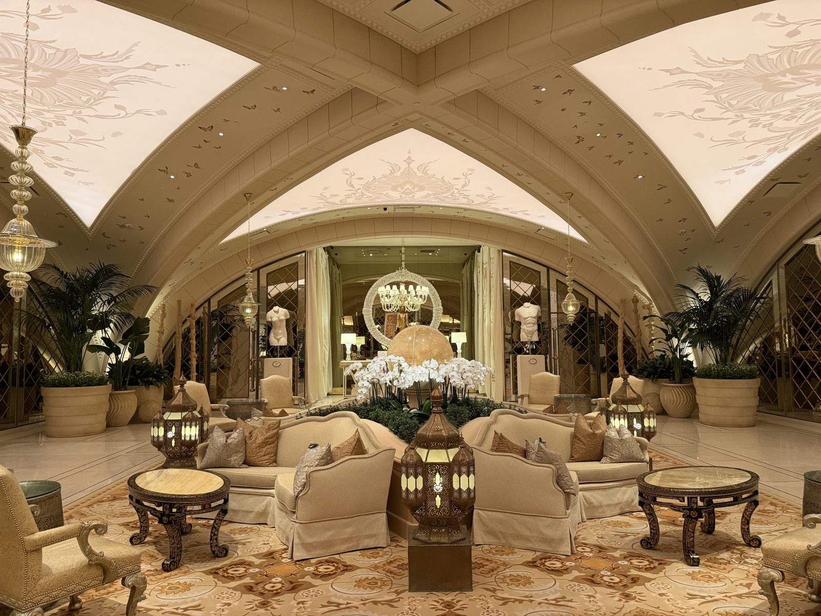 The reception lounge at The Spa at Encore Las Vegas: a cream vaulted ceiling lit from within and scattered with gilded butterflies, hanging glass lanterns, a bank of white orchids, and upholstered seating on a patterned carpet