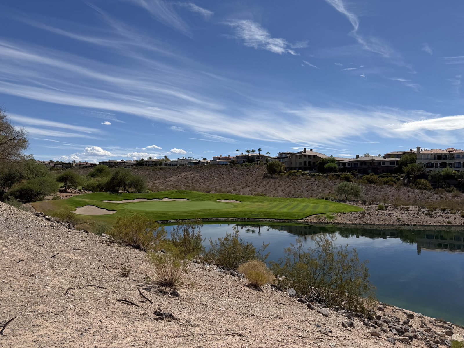 A putting green beside a still lake in Anthem with custom homes on the ridge behind