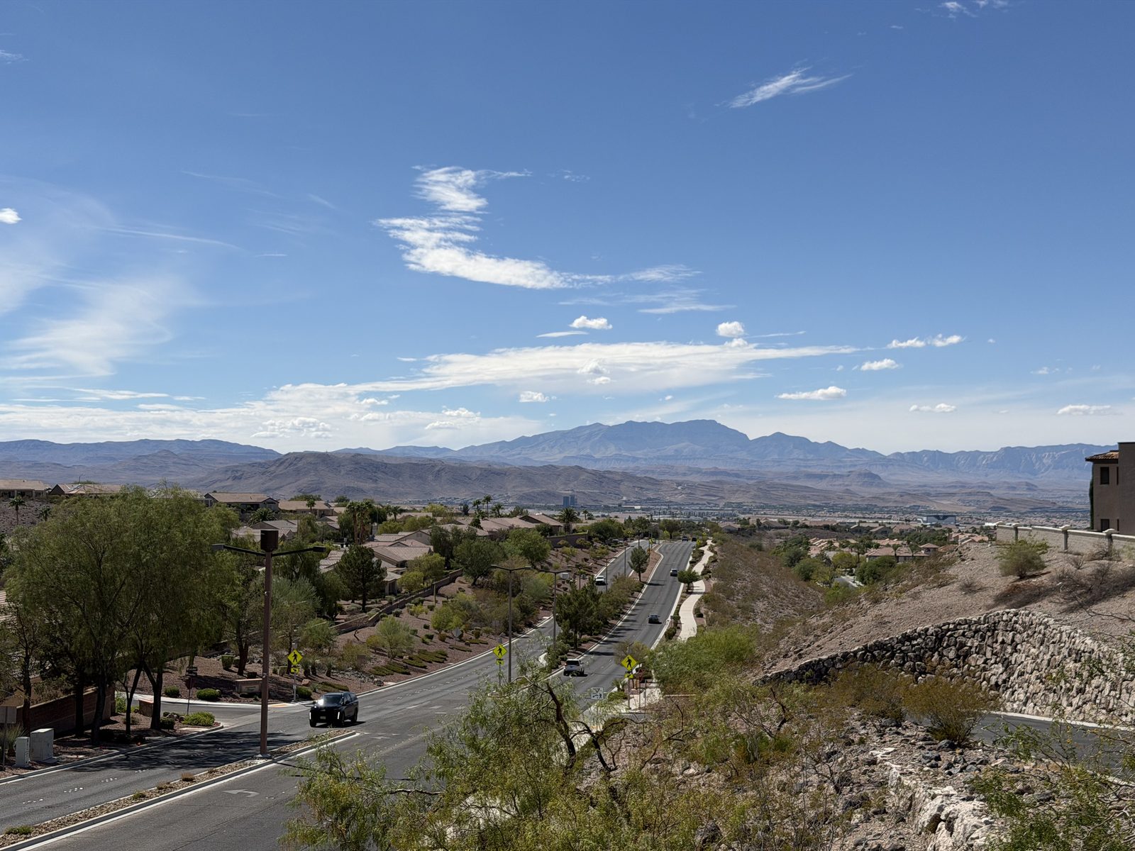 An elevated view from the Anthem hillside in Henderson, Nevada, looking down over tile-roofed homes and a descending road toward the valley floor and the mountain ranges beyond