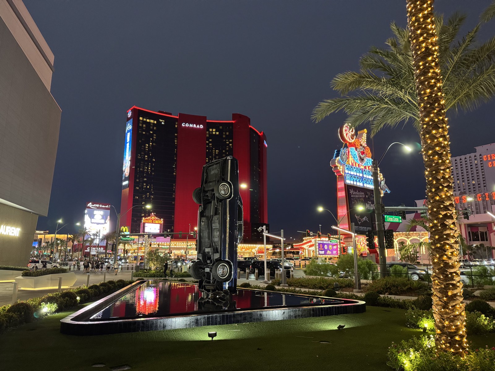 The Resorts World towers and the neon Vegas sign on the north Las Vegas Strip at night, with a vertical car sculpture in a reflecting pool in the foreground